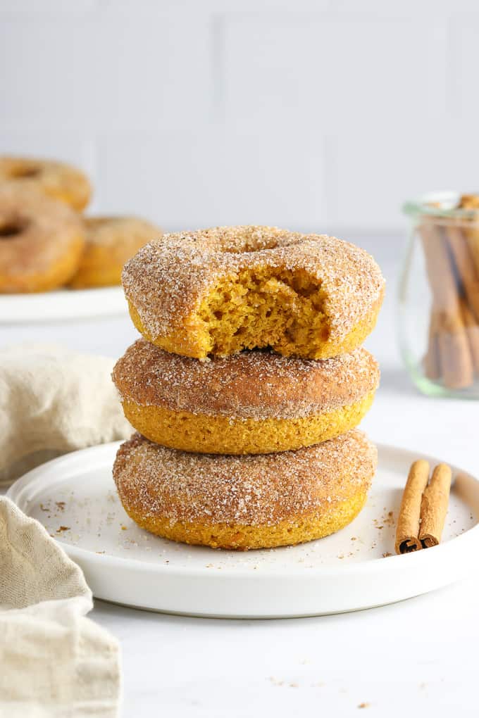 Three vegan pumpkin donuts on a white plate with cinnamon sticks and donuts in the background. 