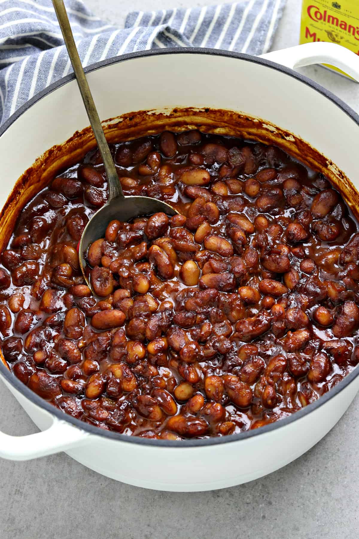 Fully cooked beans in a white pot with a spoon in the middle Napkin and mustard powder in the background.