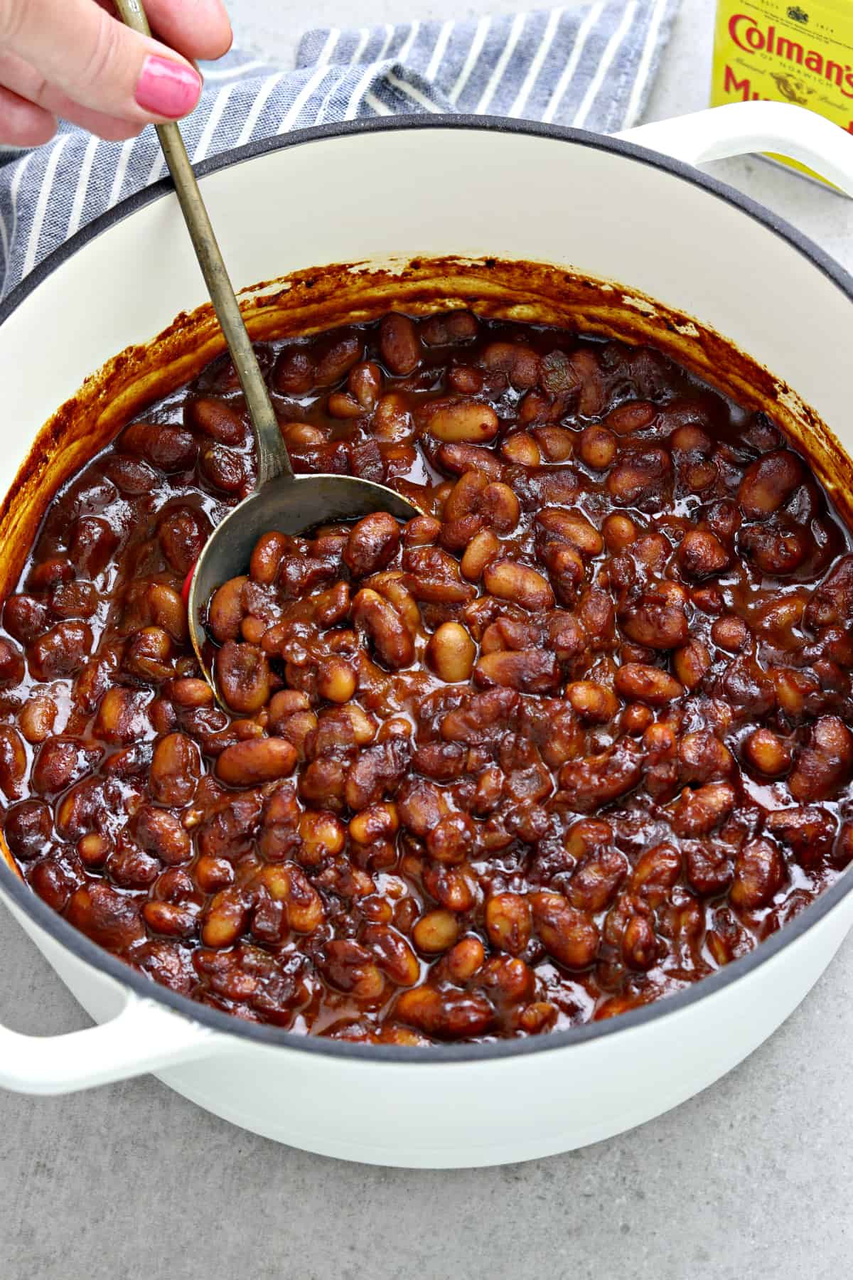 overhead view of baked beans in a white pot. Hand holding spoon and stirring beans.