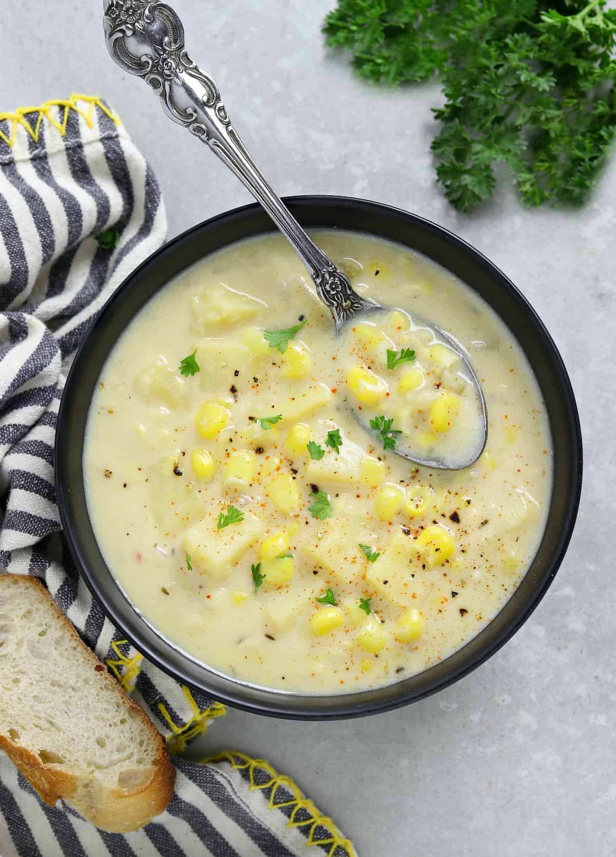 Overhead view of corn chowder in a black bowl with a spoon inside. Bread and parsley on the side.