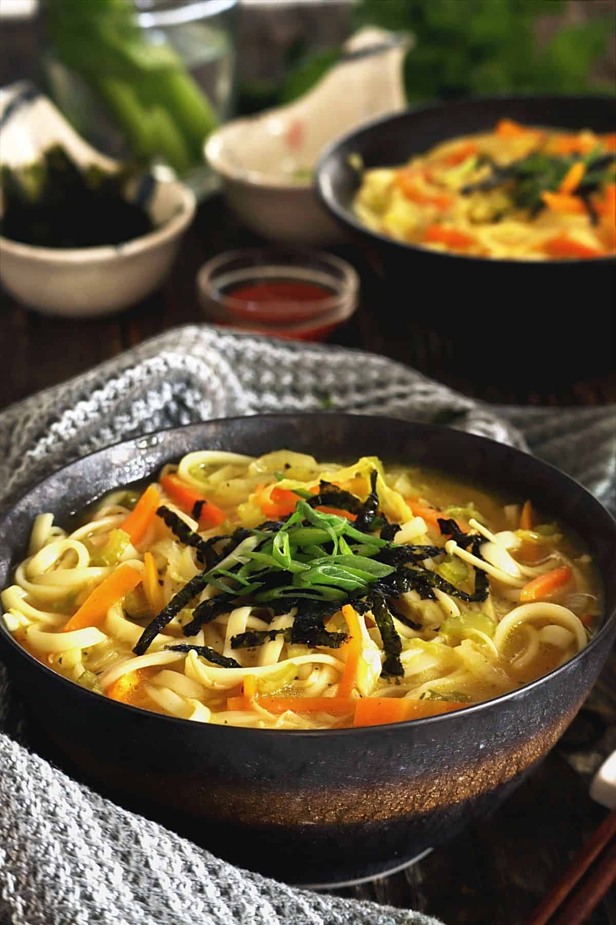 Two bowls of udon noodle soup on a wood table with sauce and nori in the background.