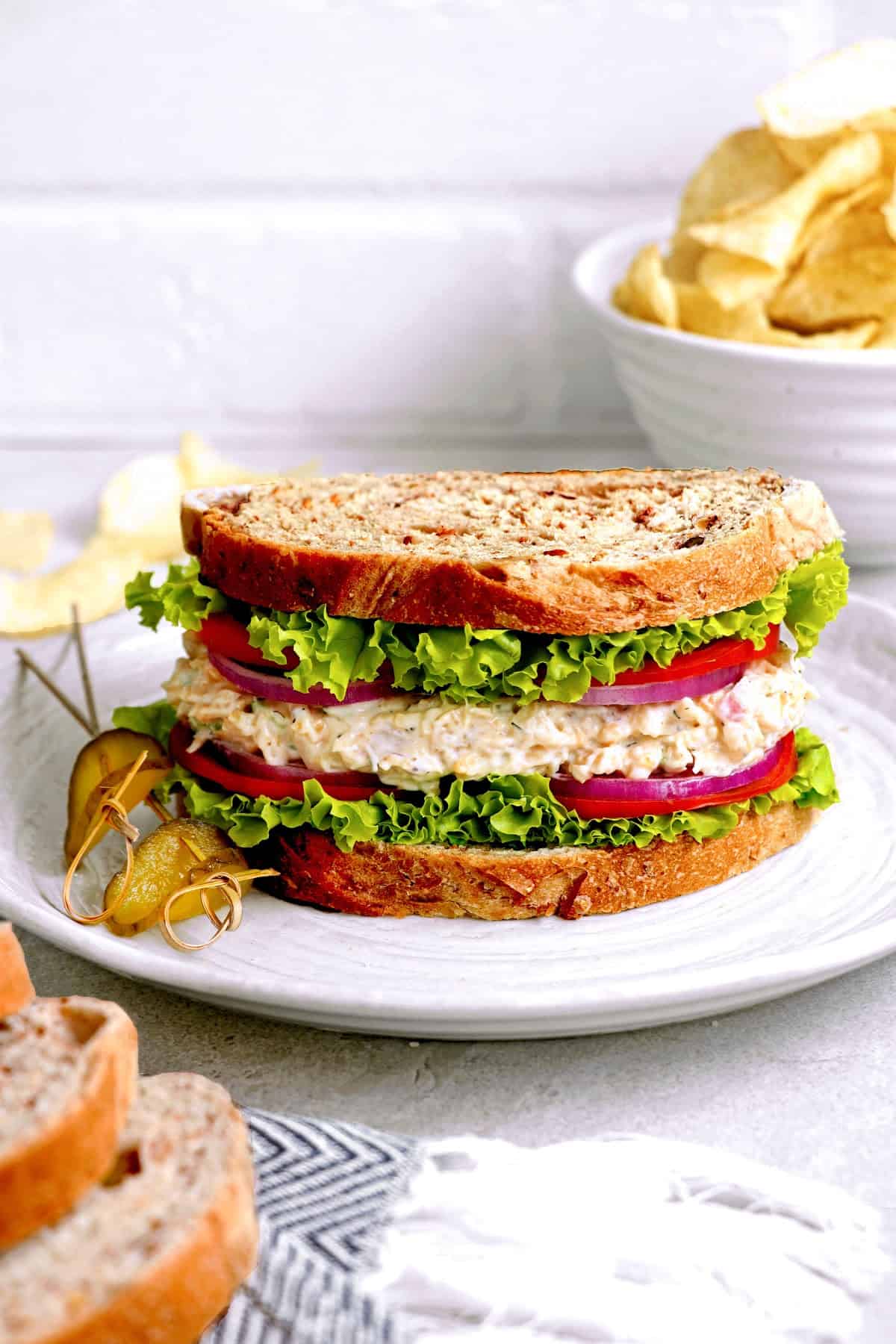 Front view of vegan tuna sandwich on a white plate. Potato chips in the background.