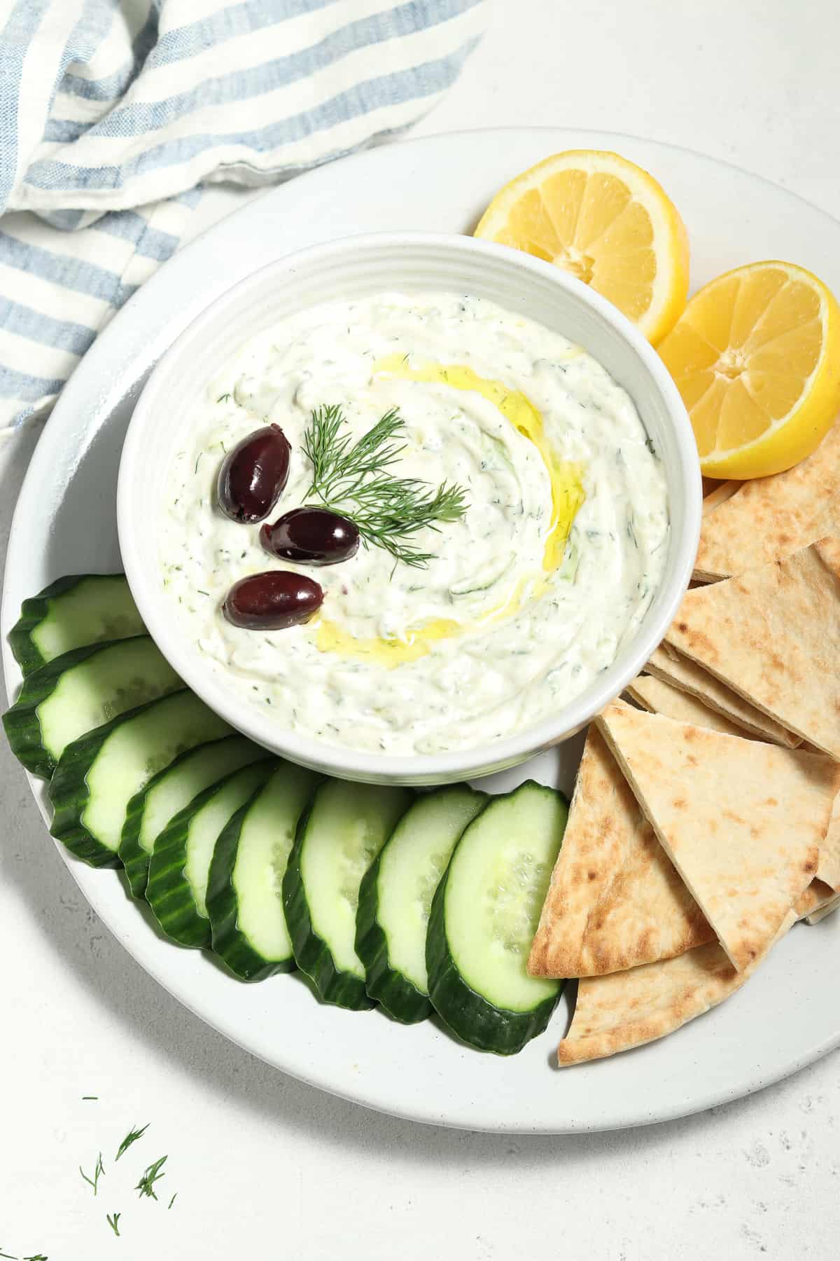 Overhead view of vegan tzatziki sauce in white bowl surrounded by pita slices and cucumber slices. 