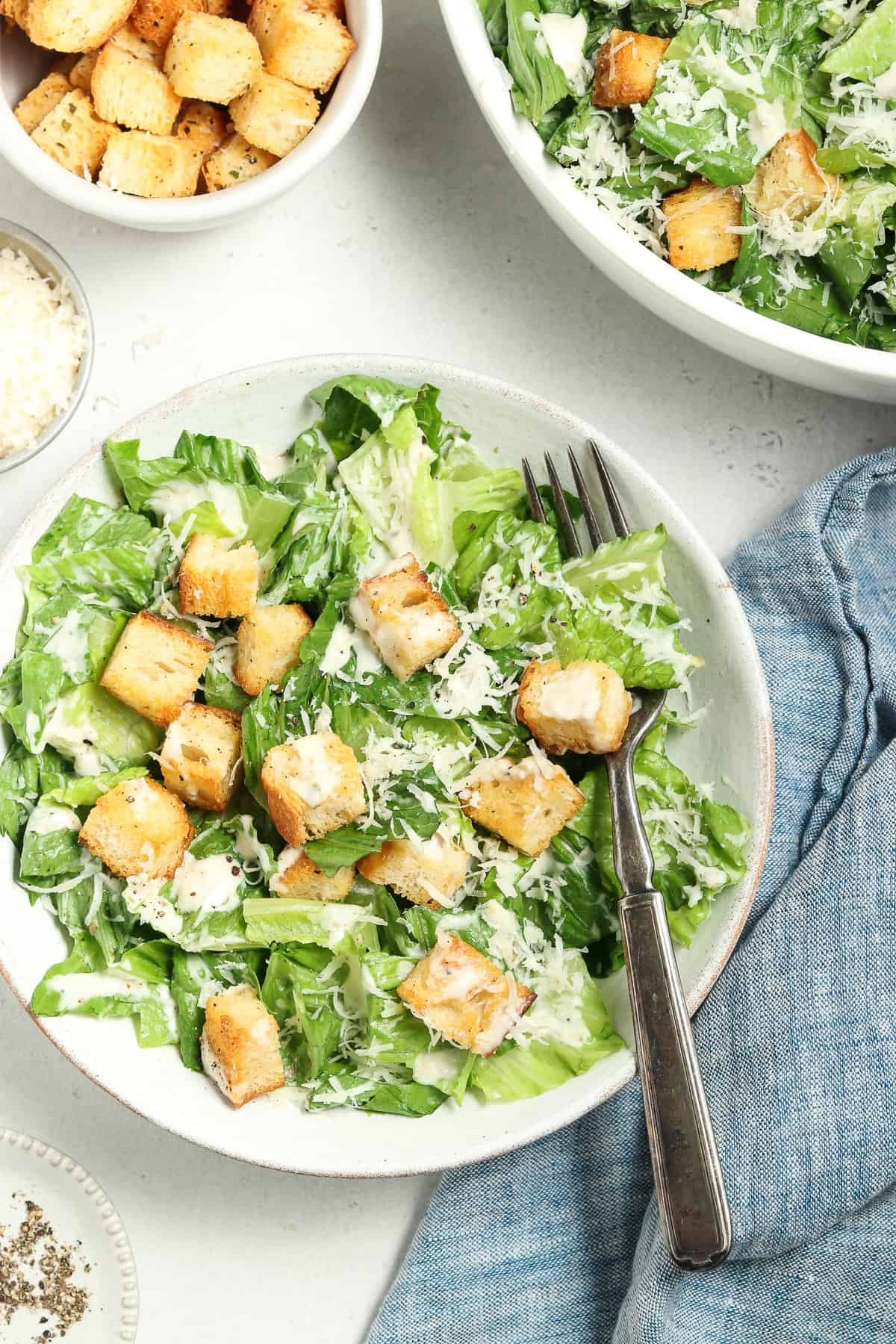 Overhead view of a vegan caesar salad in a white bowl with a blue napkin, pepper and croutons on the side. 