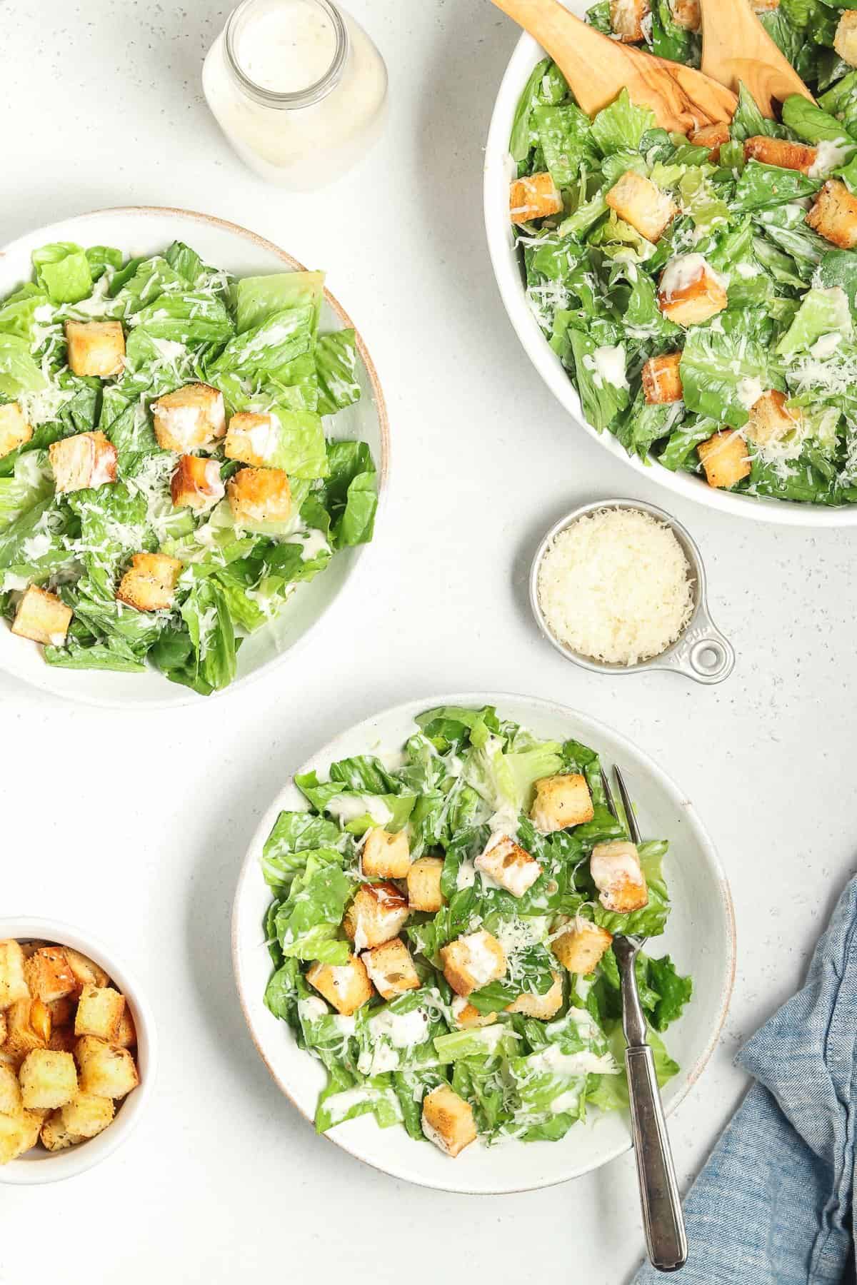 Full overhead view of 3 bowls filled with vegan caesar salad. Croutons, blue napkin and parmesan on the side.