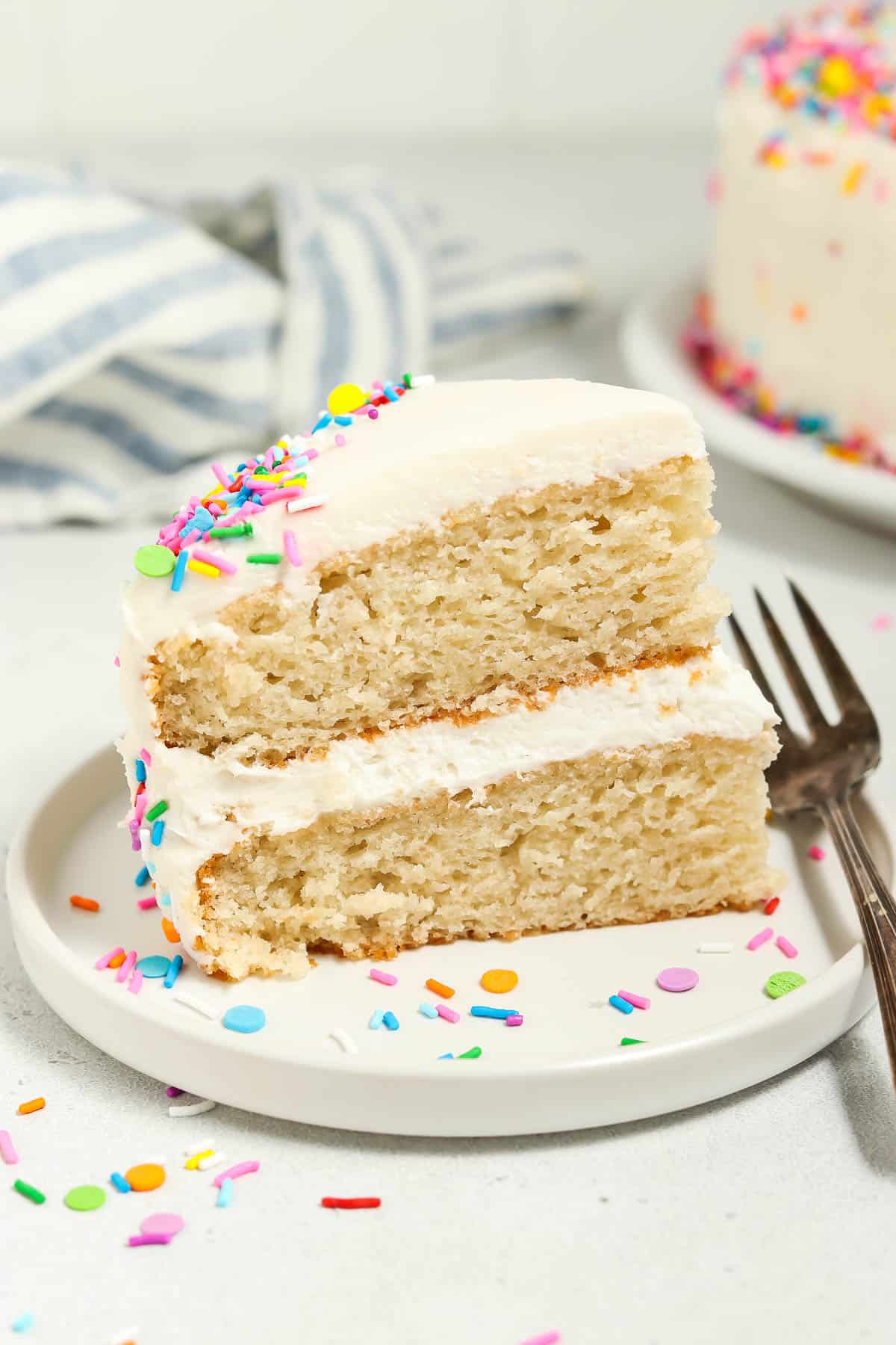 Side view of a slice of vegan vanilla cake on a white plate with sprinkles and fork on the side. 