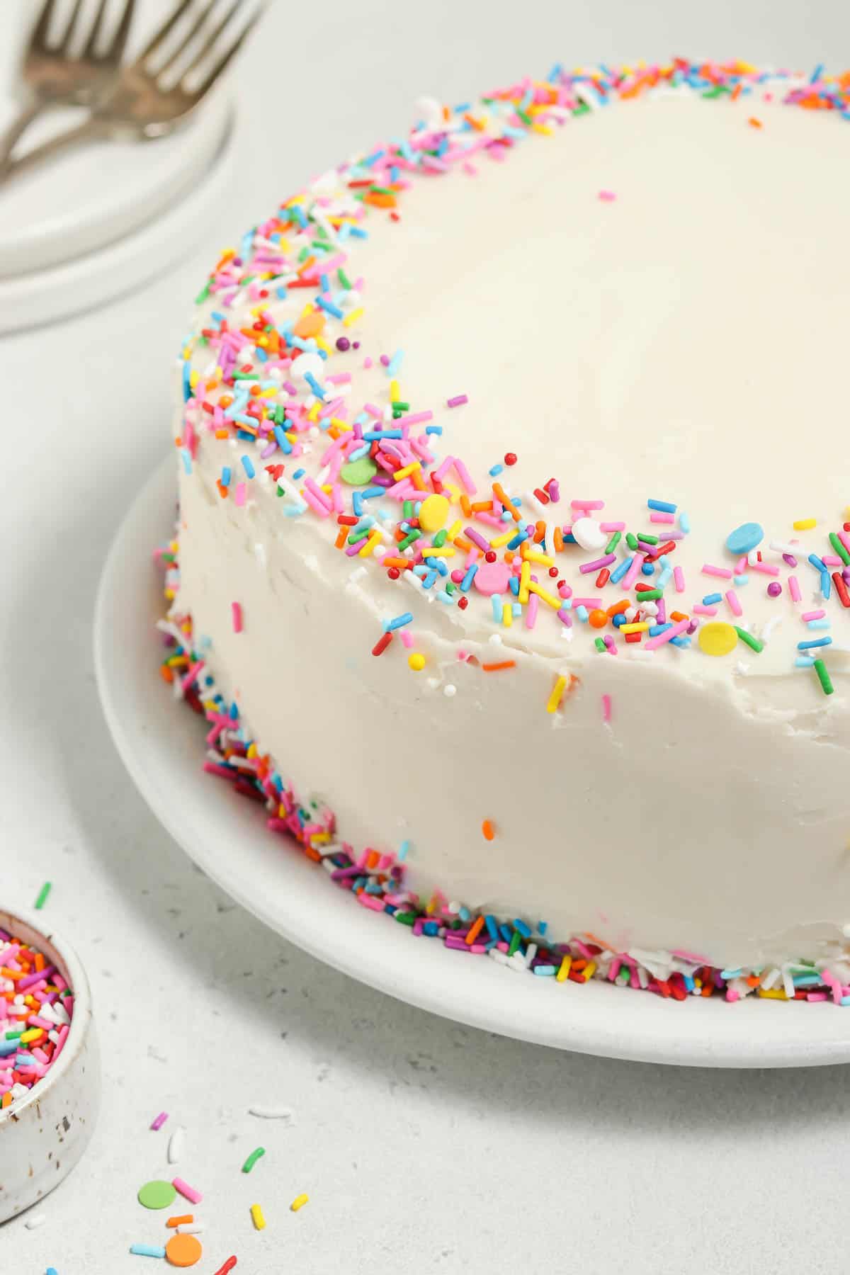 Front view of a whole vegan vanilla cake on a cake plate. Sprinkles, plates and forks on the side. 