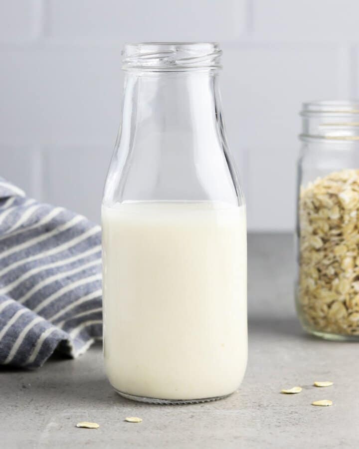front view of plant milk in a glass bottle with a jar of oats in the background.