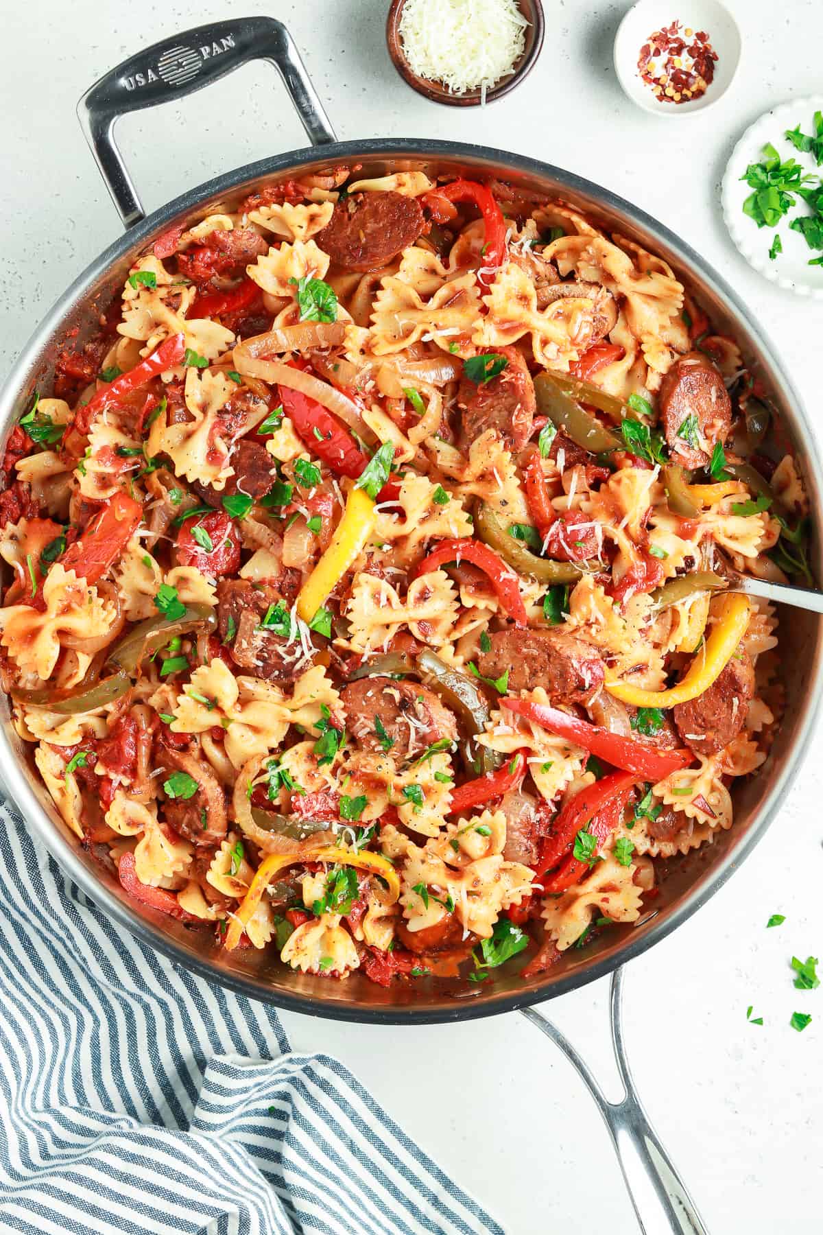 Overhead view of fully cooked sausage and peppers pasta in a pan. Parsley and parmesan on the side. 