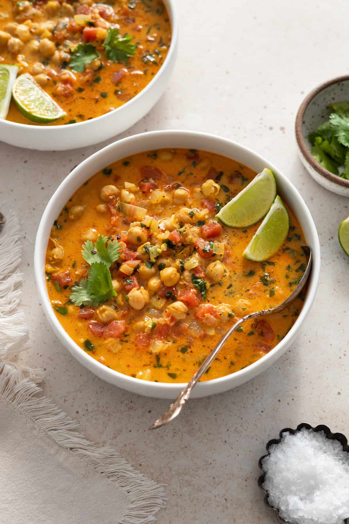 two bowls of chickpea curry soup with lime wedges and chopped cilantro on the side. 