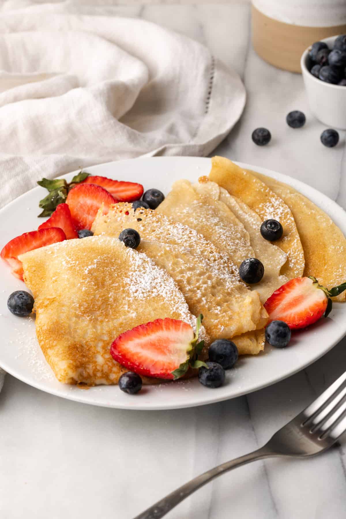 Vegan crepes on a white plate topped with powdered sugar. 