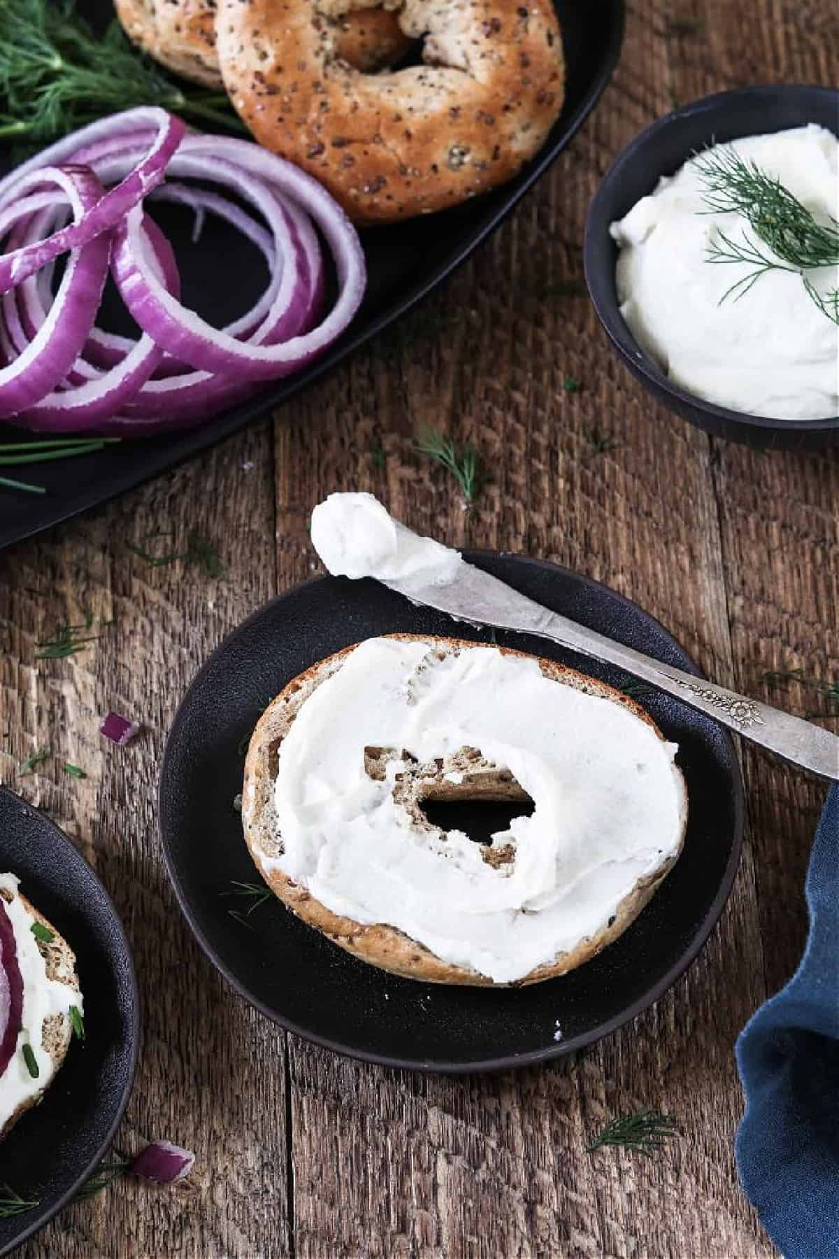 Bagel on a small black plate with vegan cream cheese, capers and dill. 