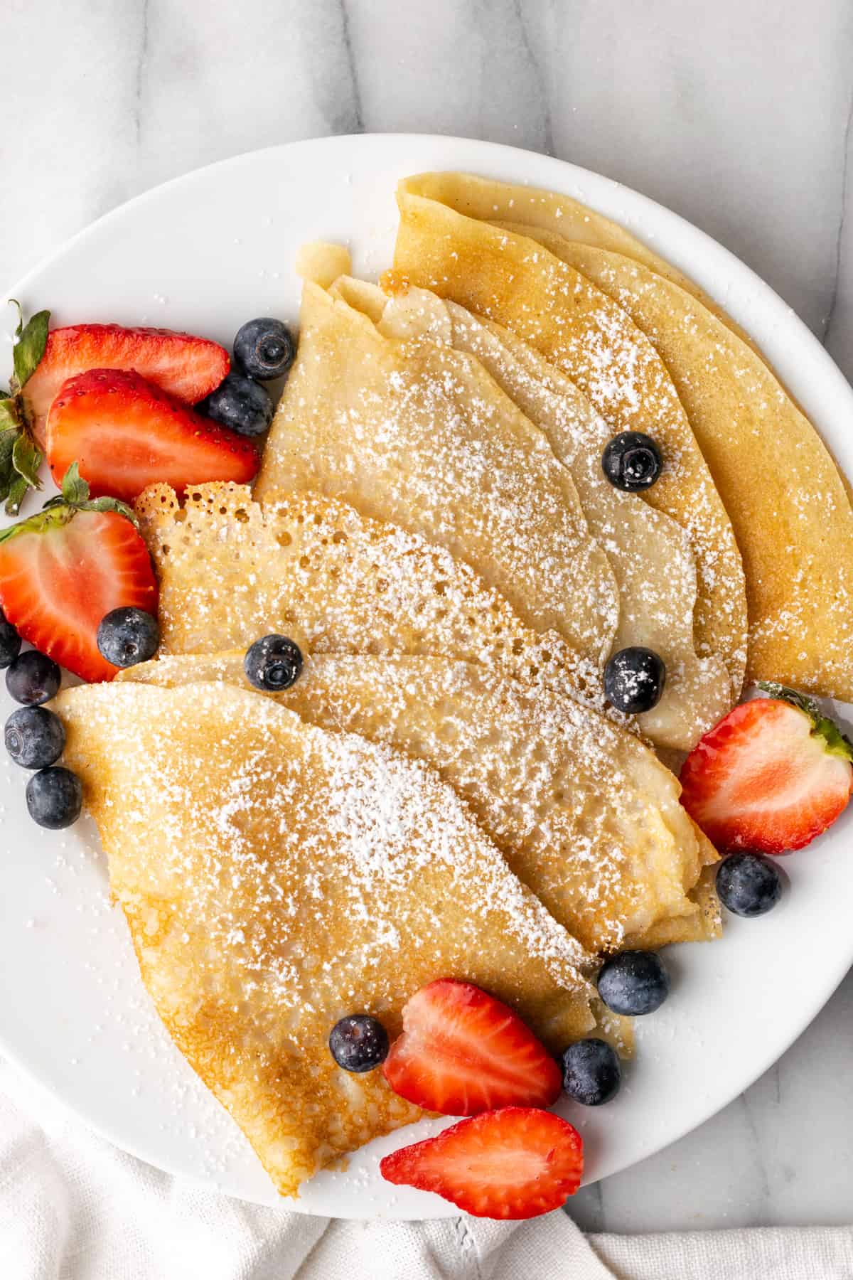 Overhead view of vegan crepes on a plate topped with powdered sugar and berries. 