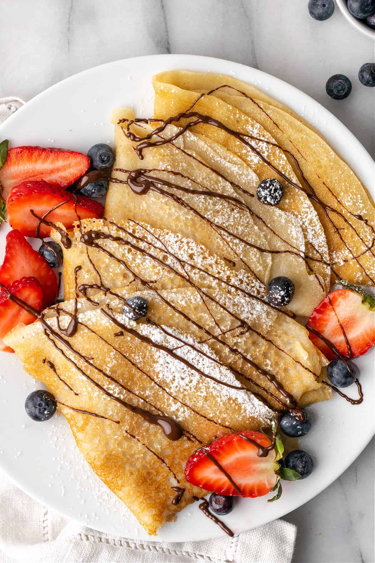 Overhead view of vegan crepes drizzled with chocolate on a white serving plate. 