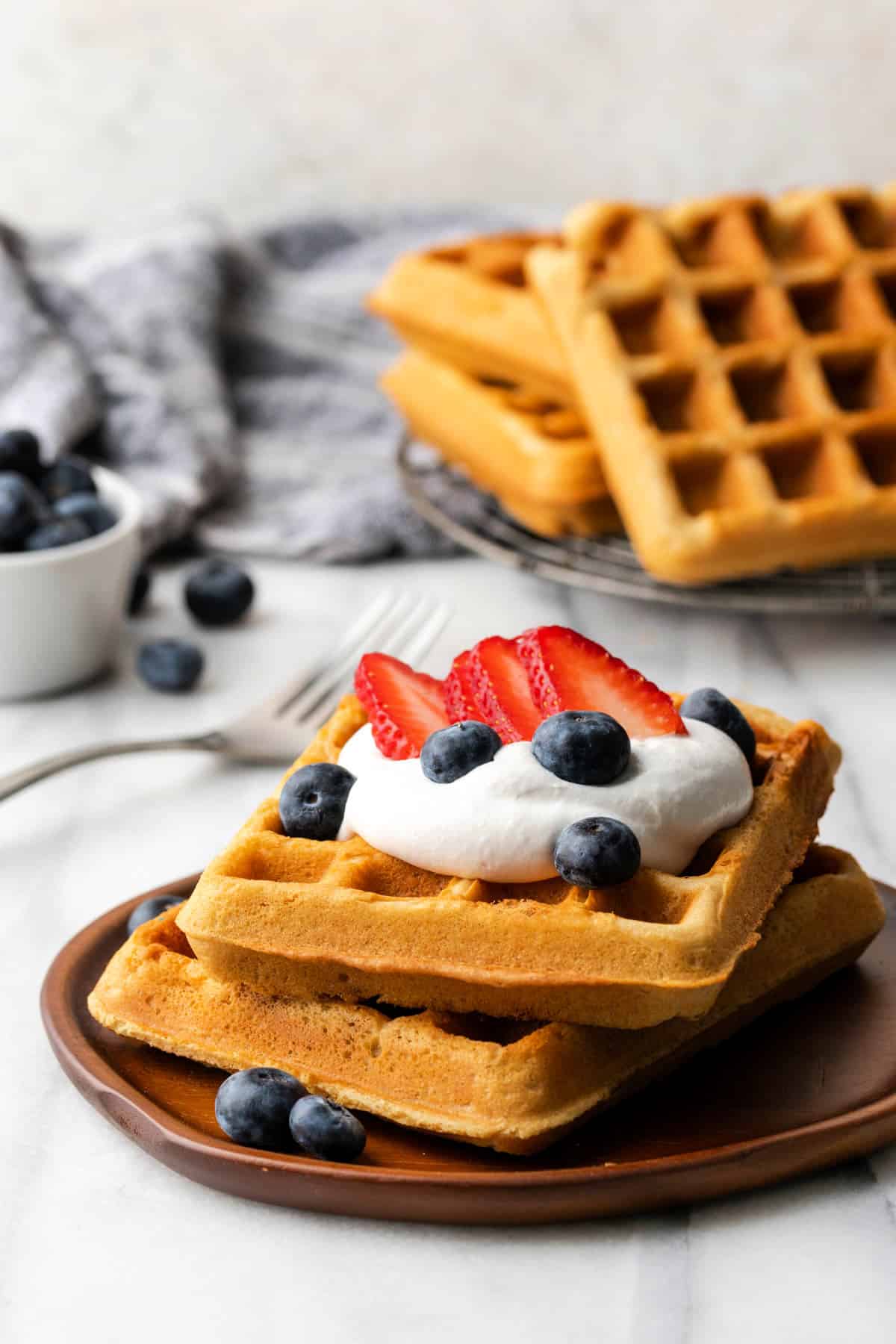Front view of two waffles on a plate with a fork and berries in the background. 
