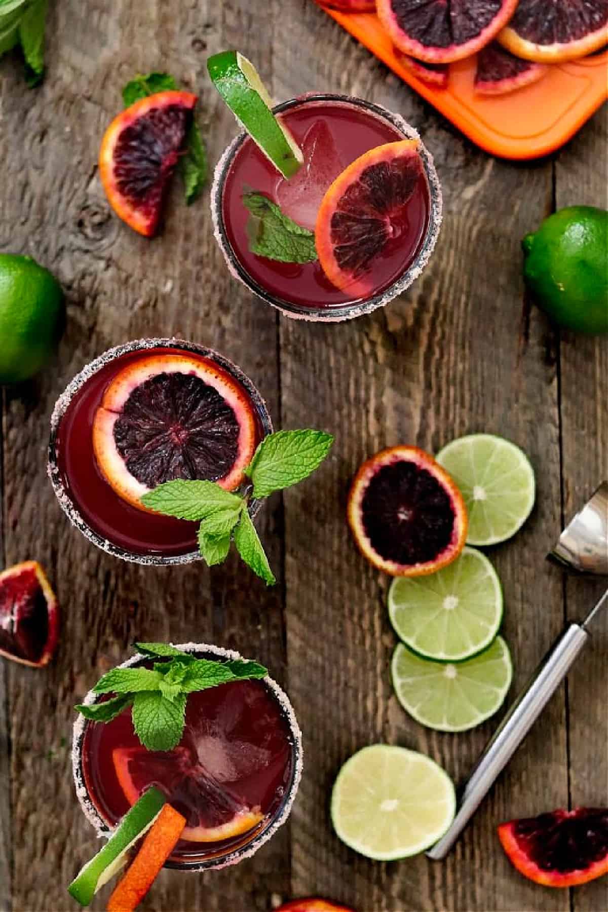 Three blood orange margaritas on a wood table top with lime and oranges on the side. 