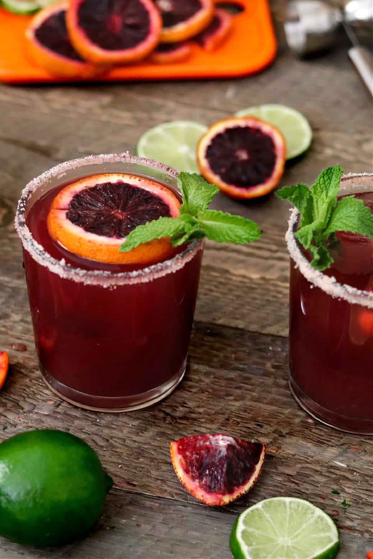 A blood orange margarita on a wooden table with lime and oranges in the background. 