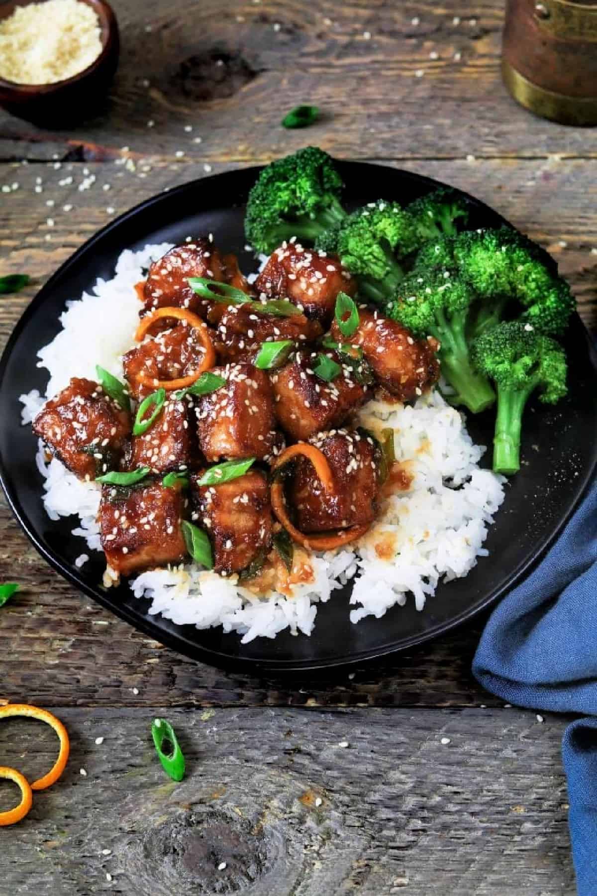 General Tsos Tofu on a black plate on top of rice and with broccoli on the side. 