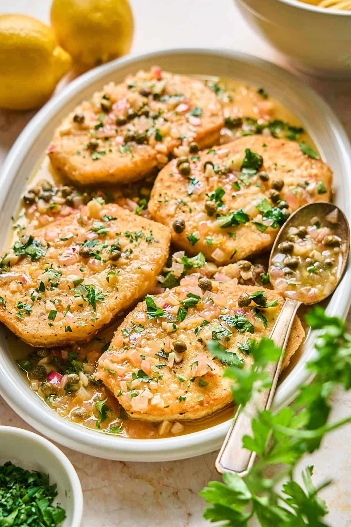 Slices of tofu piccata on a serving tray with a spoon on the side. 